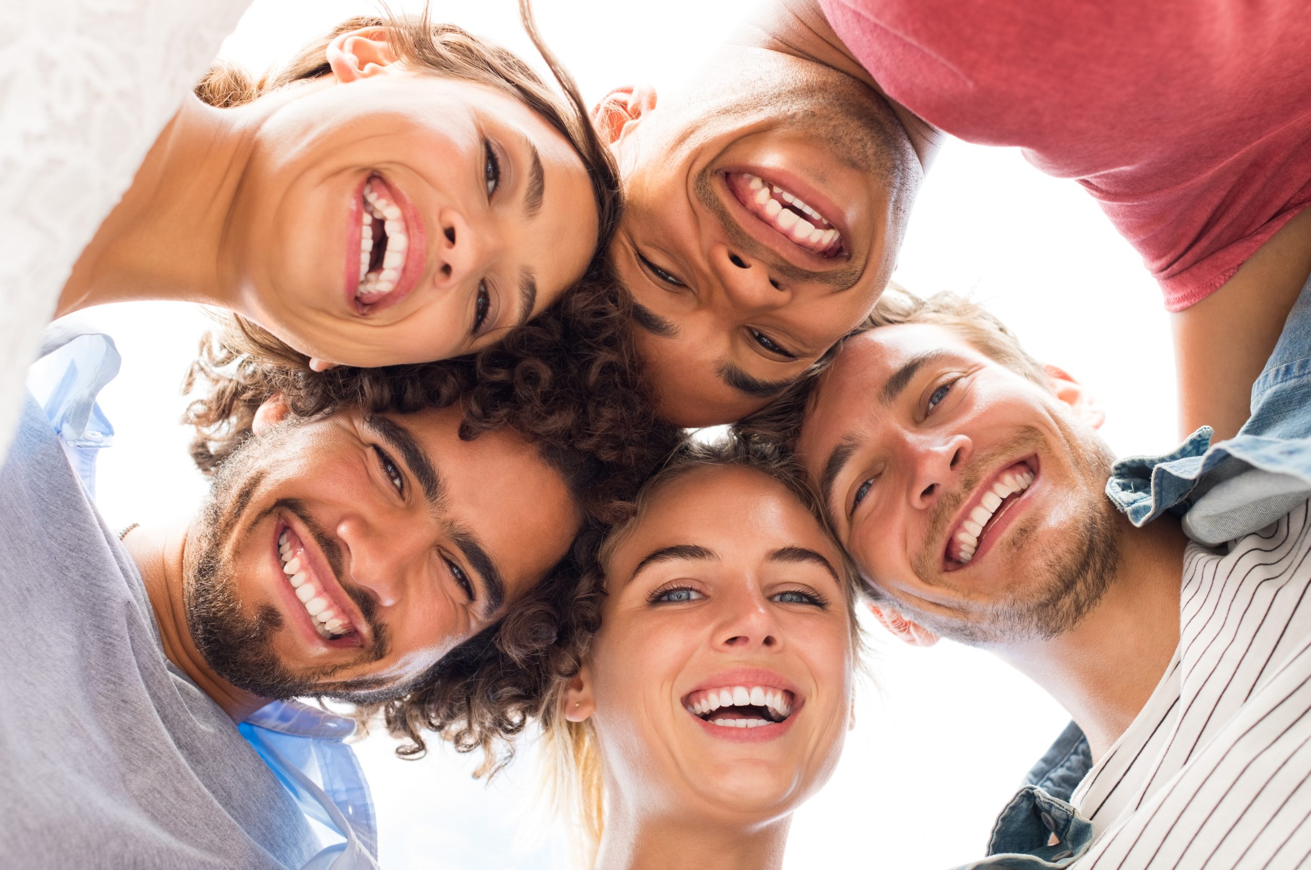Directly below shot of young friends forming huddle. Low angle view of girls and guys with their head forming a circle. Portrait of young people looking at camera. Friendship and unity concept.